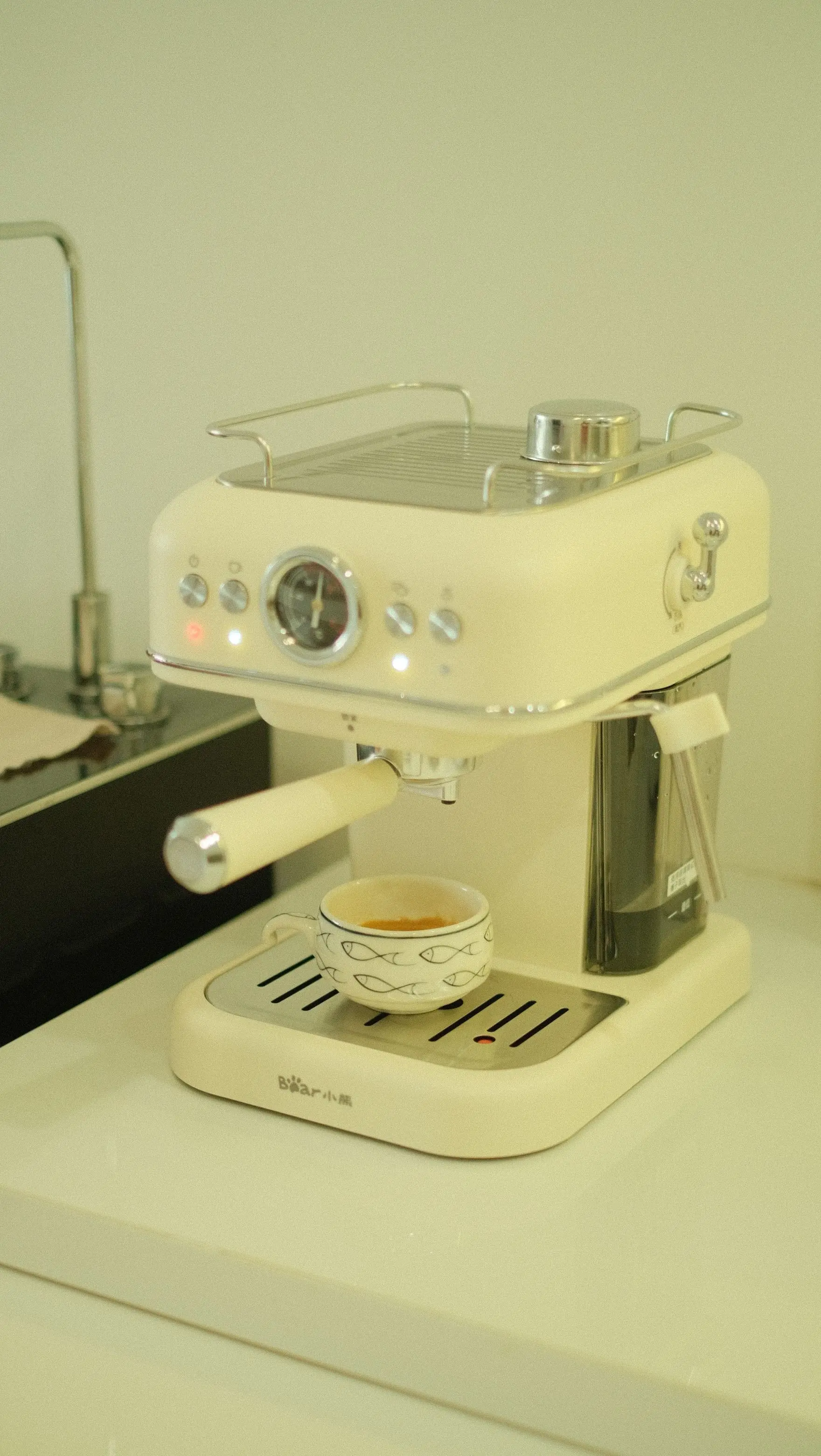 A cream-coloured espresso machine with a round pressure gauge on a white counter, a small cup of espresso on its drip tray.