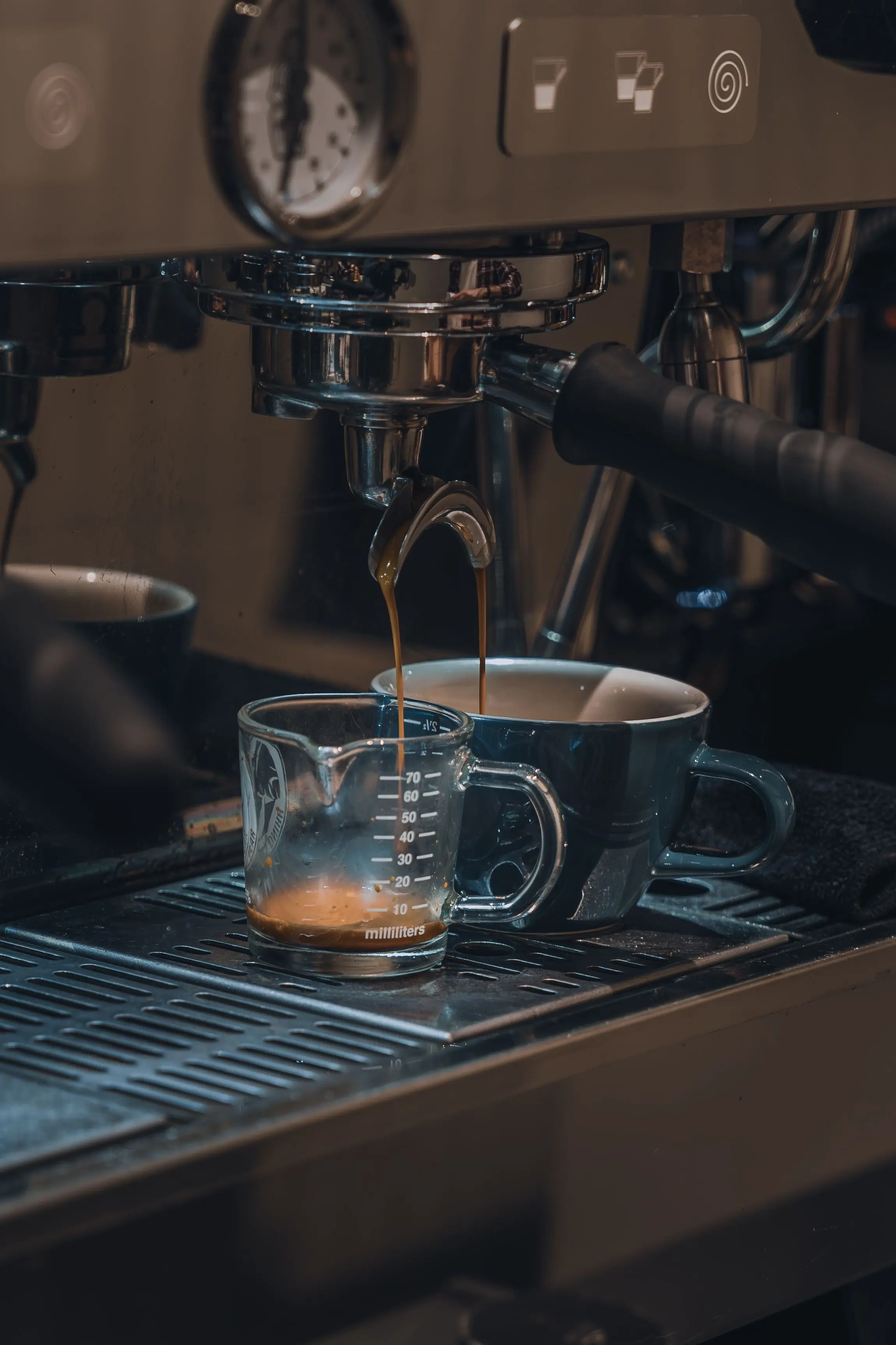 Espresso running from a portafilter spout into a small glass measuring jug, with a blue cup beside it on the drip tray.