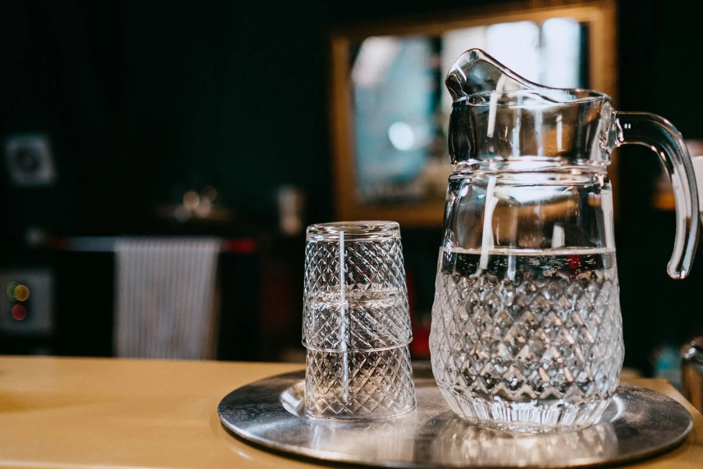 A cut glass jug of water and a matching tumbler on a metal tray, a dark room behind them.