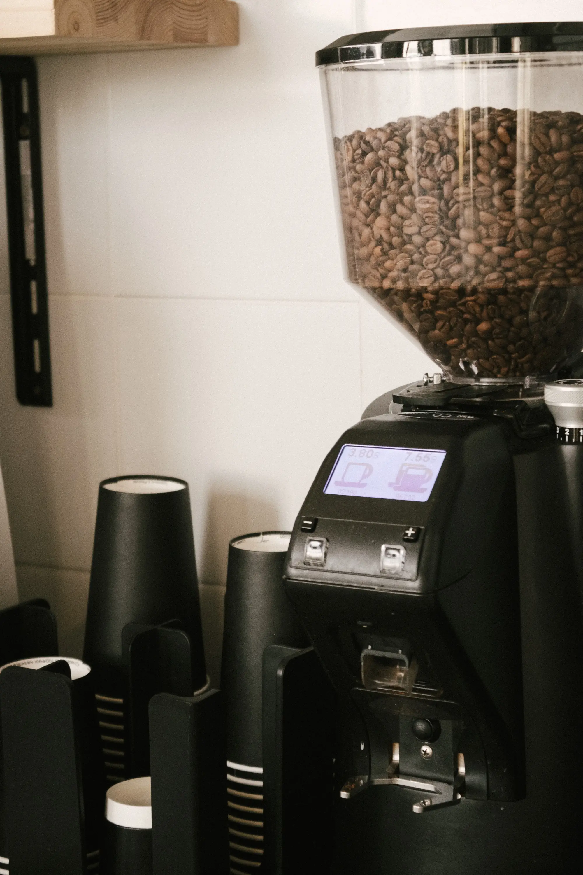 A black grinder with a clear hopper of roasted beans and a small screen, stacked cups beside it against a tiled wall.