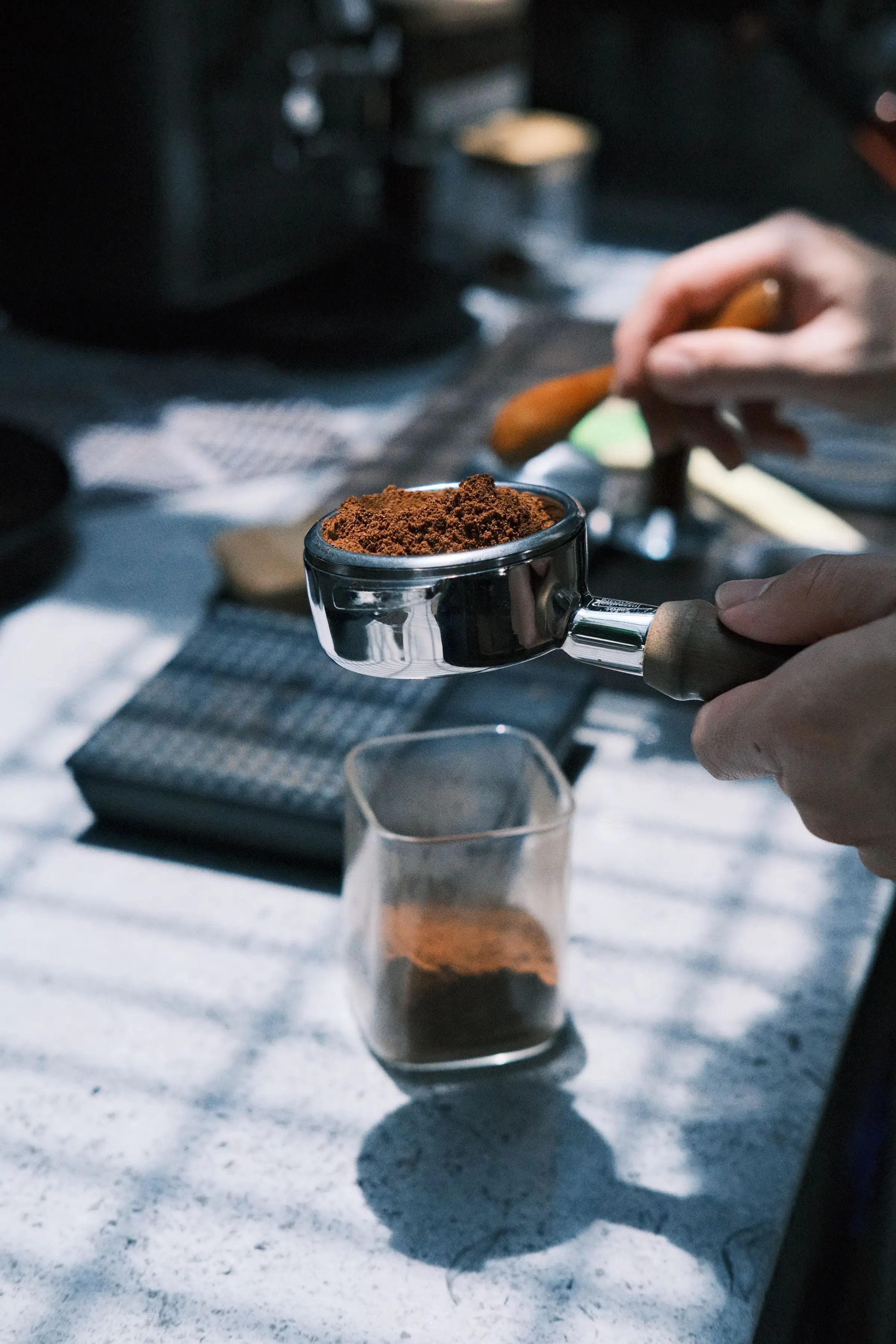 A hand holding a chrome portafilter heaped with ground coffee above a counter, a glass of grounds and a second hand behind.