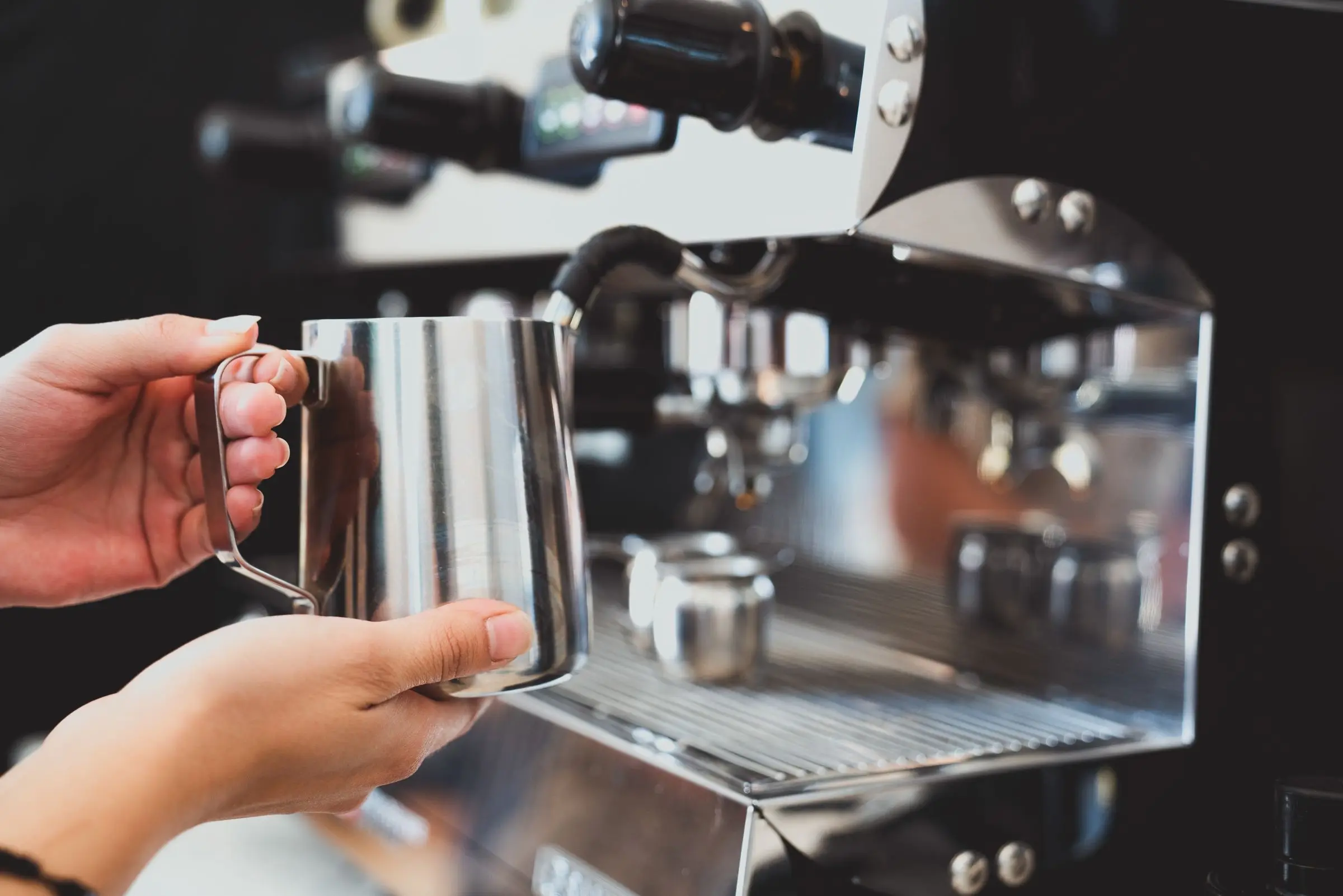 Two hands holding an empty stainless steel milk jug in front of a polished coffee machine.