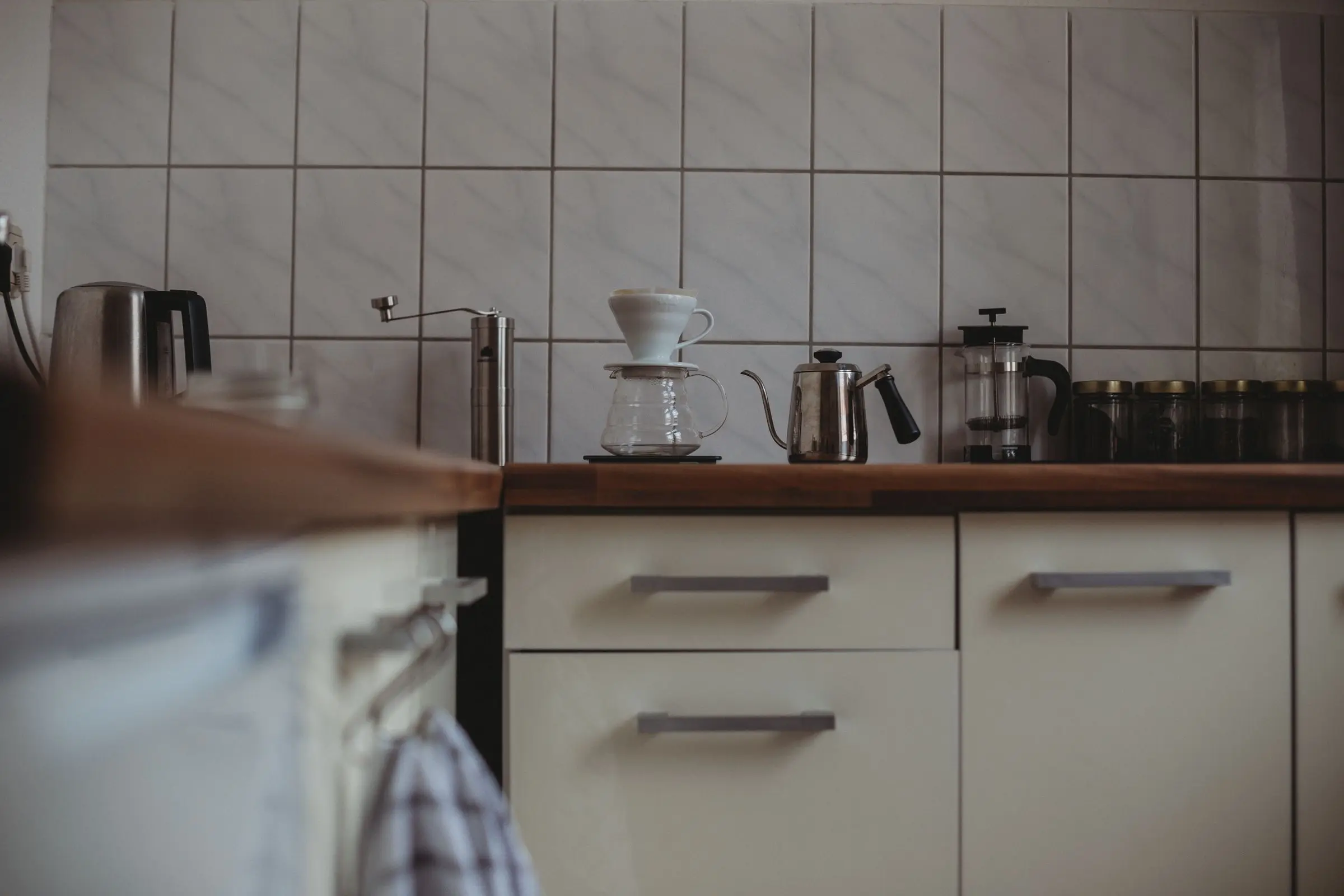 A kitchen counter with a pour-over brewer, a gooseneck kettle and a cafetiere against a tiled wall above pale cupboards.