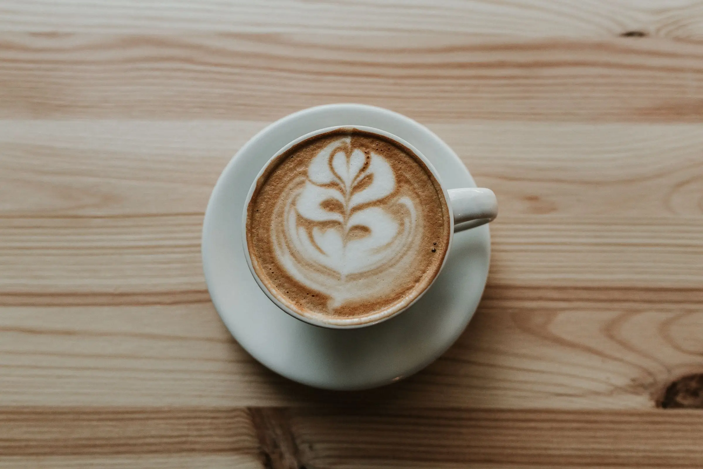 A cup of milky coffee with leaf latte art on a saucer, on a pine table, seen from above.