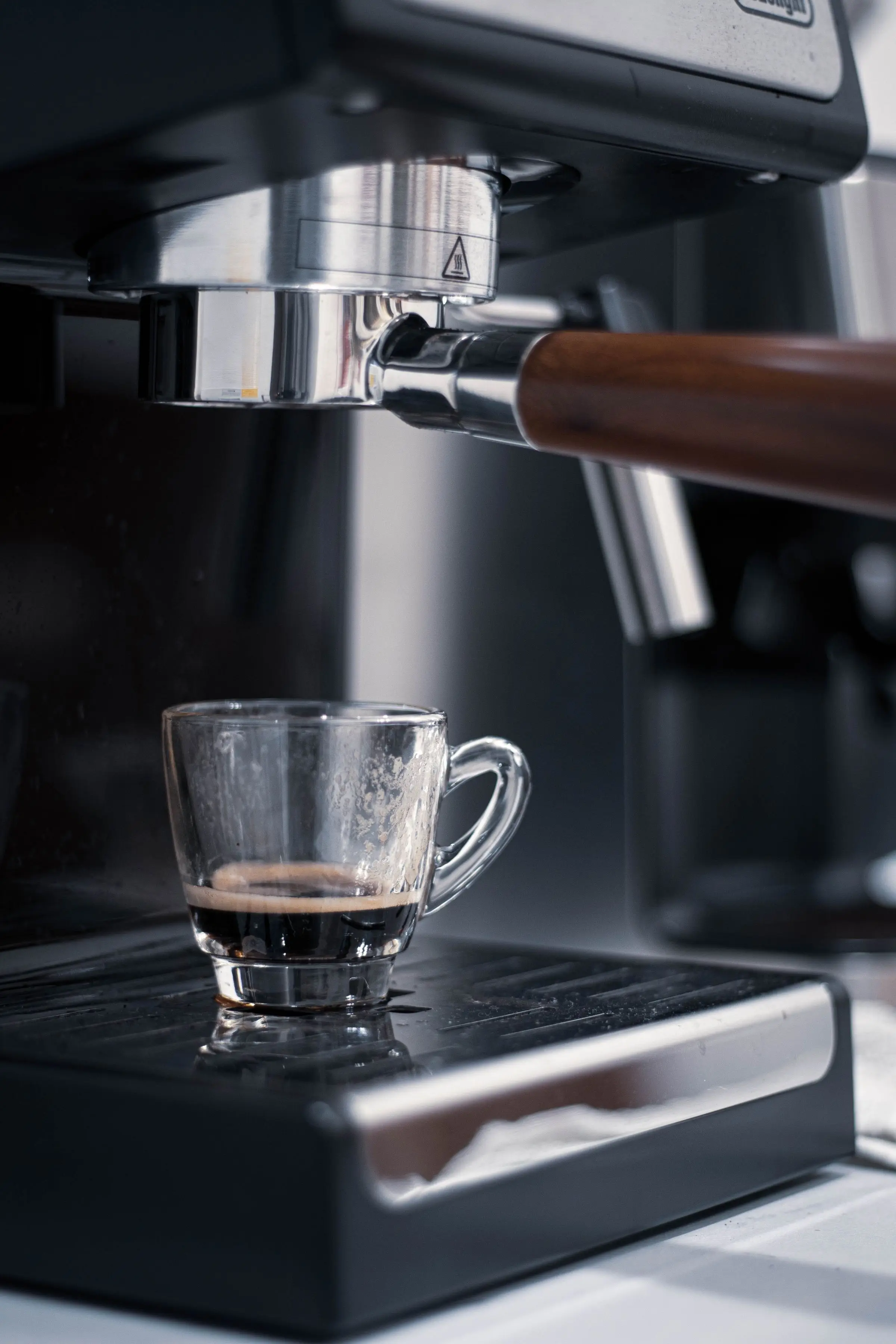 A portafilter with a wooden handle locked into the group head of a machine, a small glass of espresso underneath.