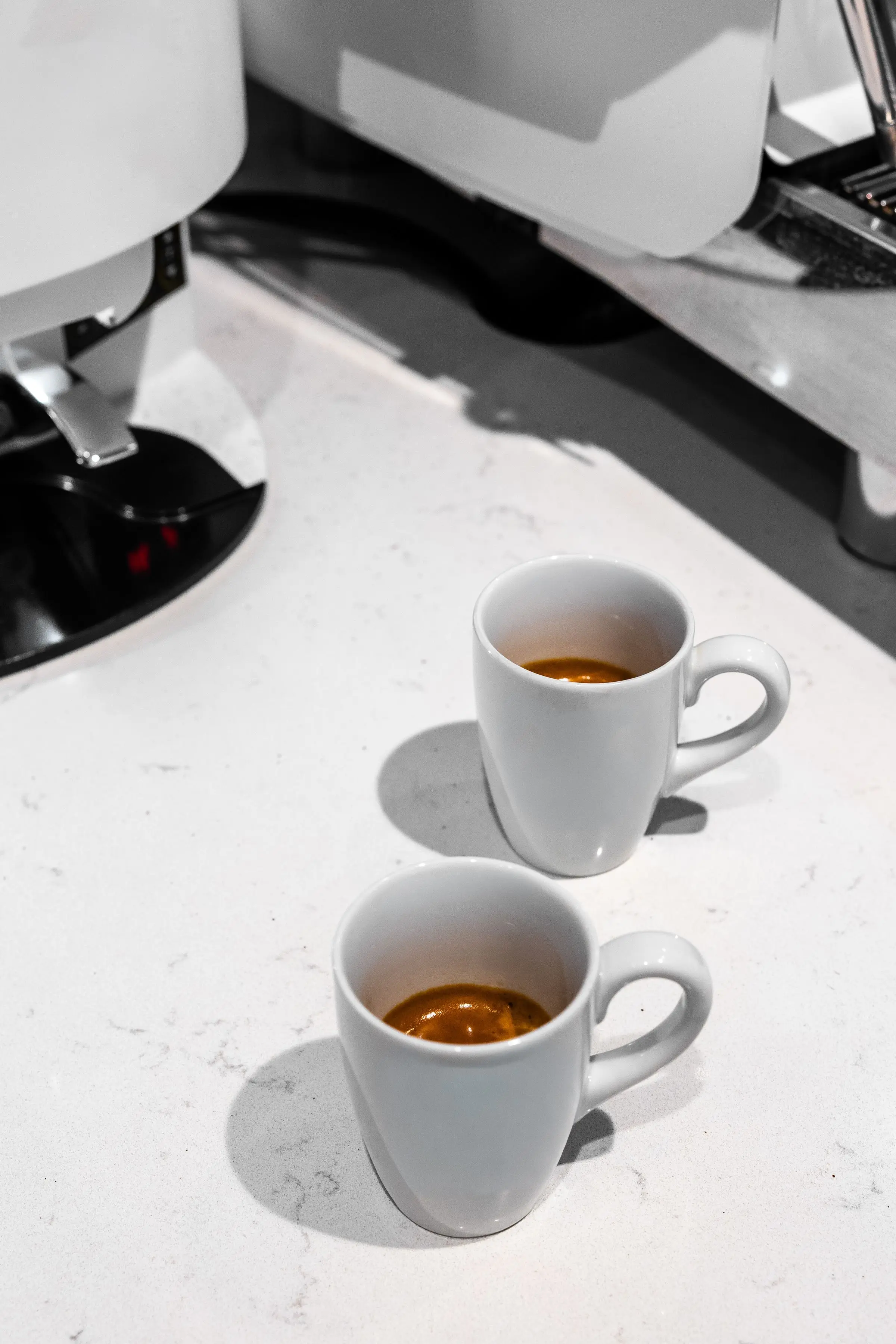 Two white cups of espresso on a white stone counter in hard sunlight, beside a grinder and a machine.