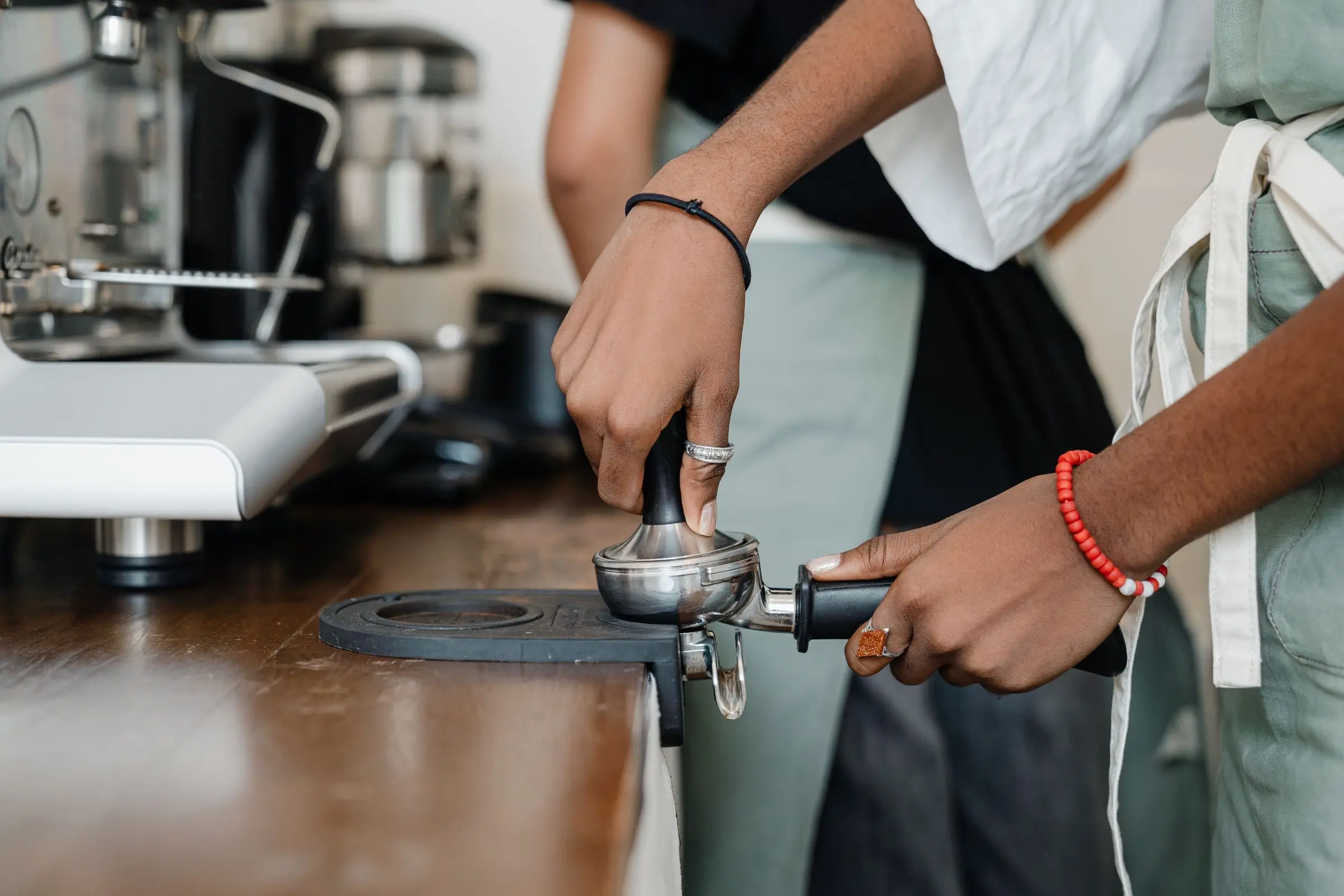 Two pairs of hands at a wooden counter, one pressing a tamper into a portafilter resting on a black mat.