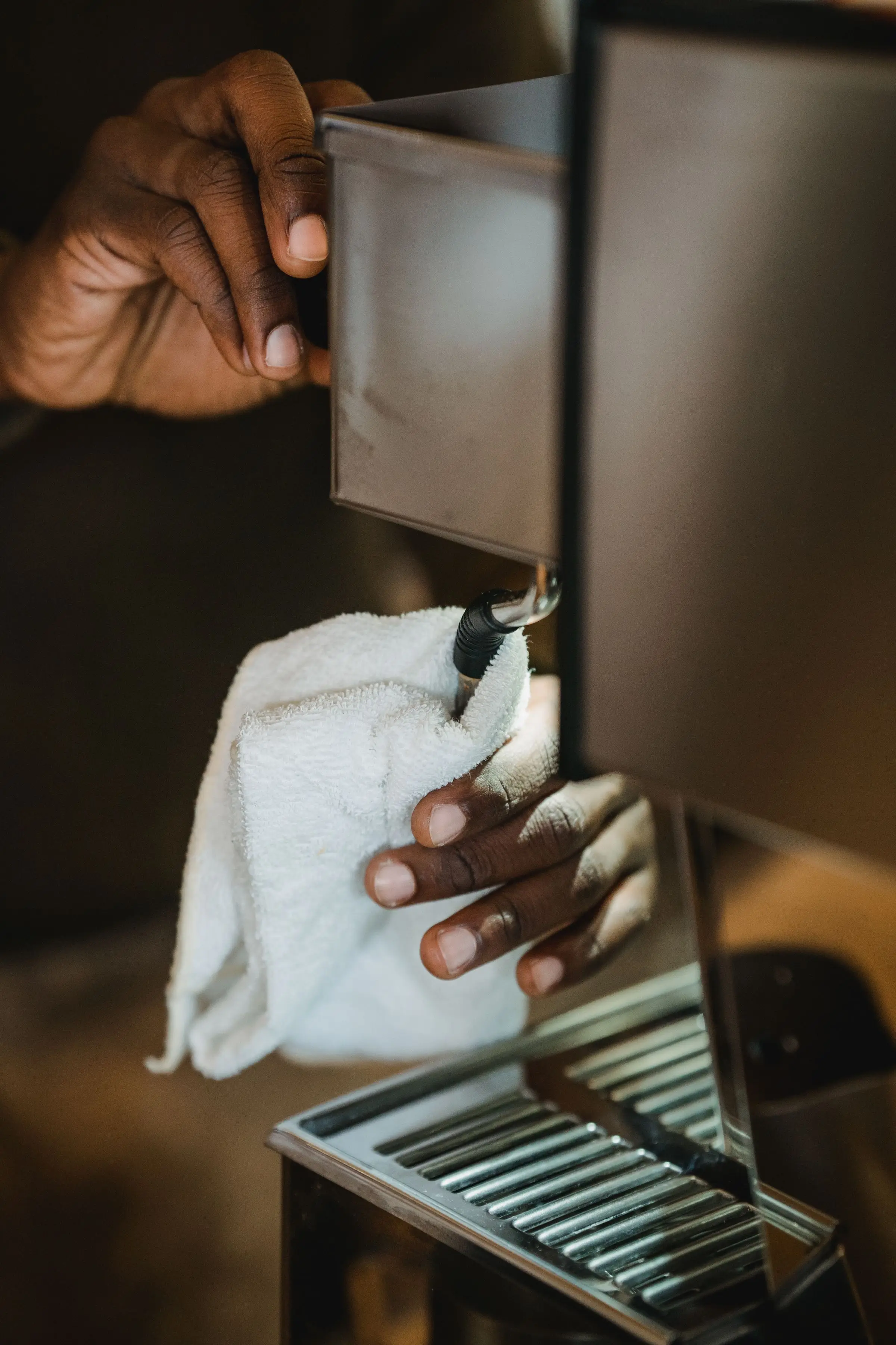 Two hands cleaning the steam wand of a stainless steel coffee machine with a white towel, the drip tray grate below.